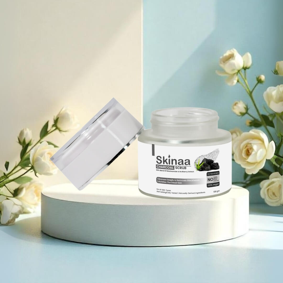 Skinaa Charcoal Scrub jar with open lid placed on a round pedestal surrounded by white roses, highlighting deep cleansing and detoxifying skincare benefits